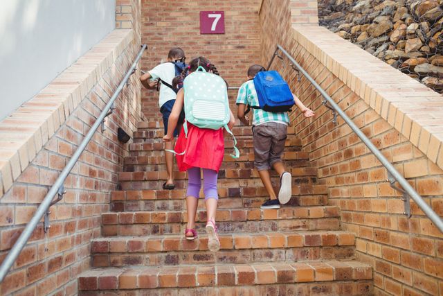 Three school kids with backpacks climbing brick stairs on a sunny day. Ideal for educational materials, back-to-school promotions, and childhood development themes. Highlights the energy and enthusiasm of young students heading to school.