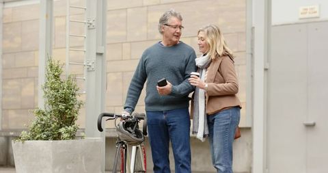 Mature couple strolling arm-in-arm with bicycle, smartphone and takeaway coffee in city
