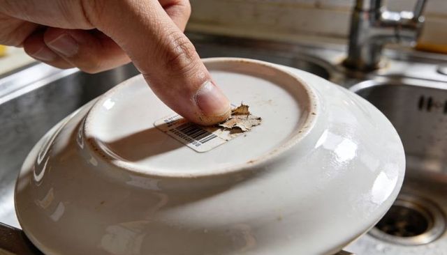 Fingertip peeling barcode sticker from ceramic plate base over stainless sink closeup