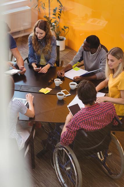 Group of diverse business professionals holding a meeting around table in modern office. Individuals are engaging in discussions, using technology and taking notes. Suitable for themes related to teamwork, business strategy, office environment, inclusion, and technology in the workplace.