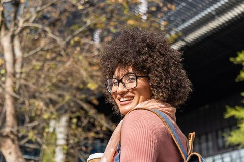 Smiling curly-haired woman holding coffee, wearing glasses and backpack in sunlit city plaza