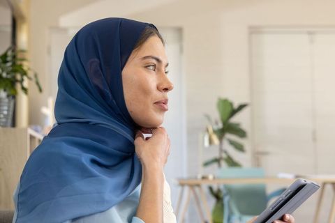 Thoughtful muslim woman wearing blue hijab holding smartphone and touching throat at home