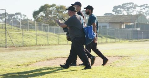 Diverse teammates walking across baseball field carrying gear and bags wearing caps