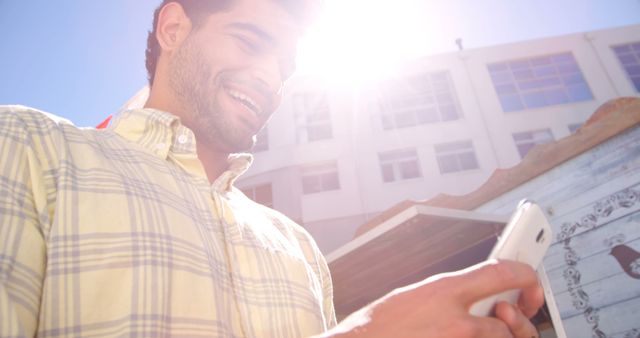 A young Caucasian man smiles as he uses his smartphone in an urban setting, with sunlight flaring in the background. His casual attire and cheerful demeanor suggest he might be enjoying a leisurely day or engaging in social communication.