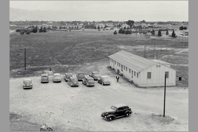 This scene offers a glimpse of a historical time period at Ames with a focus on a construction shack surrounded by vintage cars and landscape, looking toward the southwest. Still-utilized Navy housing and buildings can be seen in the background, adding context to the wartime era. This would be suitable for articles on historical architecture, 1940s American history, vintage transportation, or exhibitions on wartime life in the United States.