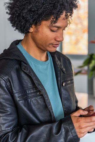 African american man wearing leather jacket checking smartphone, curly hair indoor portrait