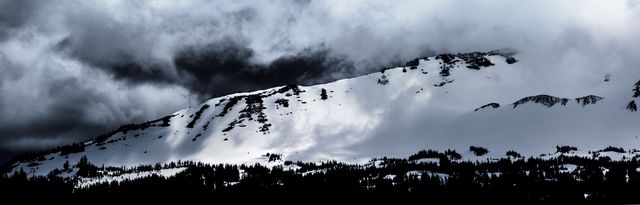 Dark storm clouds gather ominously over a snow-covered mountain range. The snowy slopes and dense forest create a dramatic and atmospheric winter scene. Ideal for use in travel promotions, weather reports, dramatic scenery collections, or nature and adventure-themed projects.