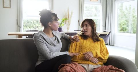Mid-adult friends chatting on cozy sofa wearing knitted sweaters in bright home living room