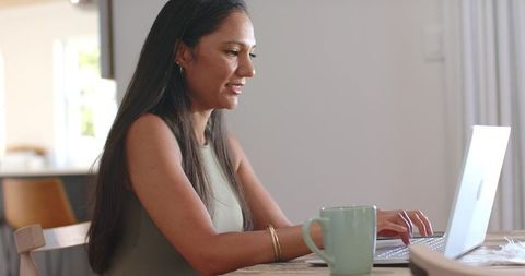 Woman typing on laptop in sunlit kitchen with coffee mug for remote work lifestyle
