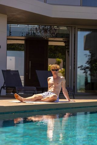 Young man sunbathing on private modern pool deck at luxury home wearing swim trunks