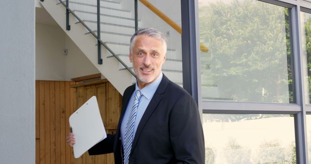 Middle-aged businessman holding clipboard, wearing suit and tie, posed near glass-walled office entrance. Suitable for use in content related to business, corporate environments, job interviews, business meetings, office concepts, or professional settings.