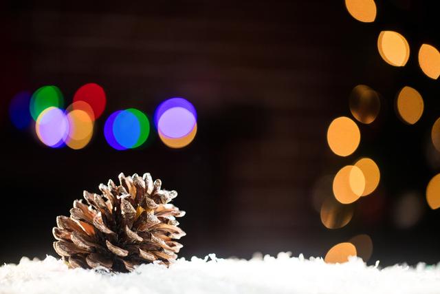 Pine cone resting on artificial snow with colorful Christmas lights in the background. Ideal for holiday greeting cards, festive decorations, winter-themed promotions, and seasonal advertisements.
