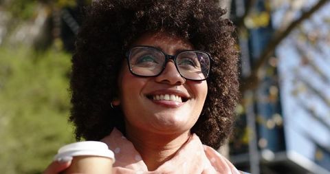 Smiling woman holding takeaway coffee cup on urban street wearing glasses and pink scarf