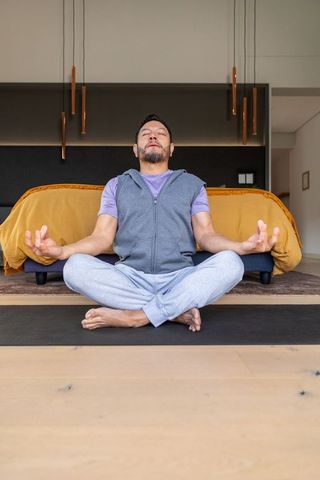 Mature asian man meditating in minimalist bedroom with mustard duvet and pendant lights