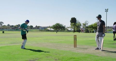 Wicketkeeper adjusting gloves while batsman waiting at crease on sunny suburban cricket pitch