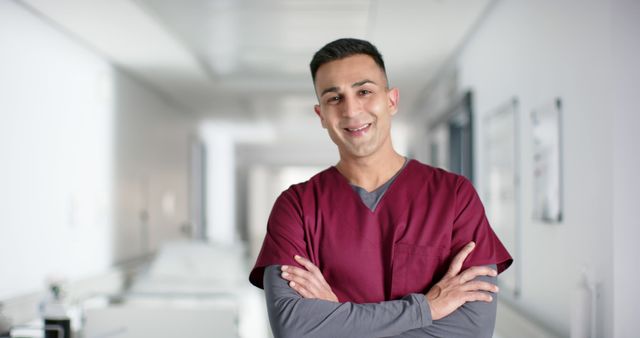 A confident healthcare professional wearing maroon scrubs stands in a bright hospital corridor, smiling with arms crossed. Suitable for topics related to healthcare, medical professionals, nurses, doctors, hospital environments, and health services. Ideal for health care promotional materials, medical training content, and health-related articles.