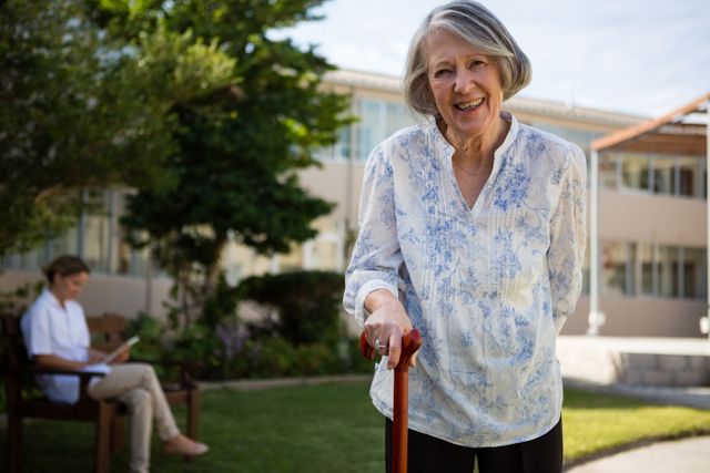 Senior woman smiling while holding a cane in a park, with a doctor sitting in the background. Ideal for use in healthcare, retirement, and wellness promotions, as well as articles on aging and senior lifestyle.