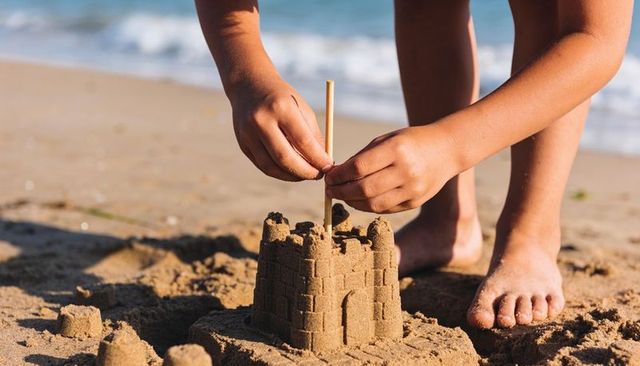 Child building detailed sandcastle on sunny shoreline placing flagpole with bare feet close-up