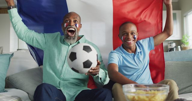Two generations of family bonding while watching a soccer match together. They are holding a French flag, showing enthusiasm and support for their team. The man is holding a soccer ball, both have face paint. Perfect for depicting family moments, sports enthusiasm, or fan culture in advertising, promotional materials, or editorial content.