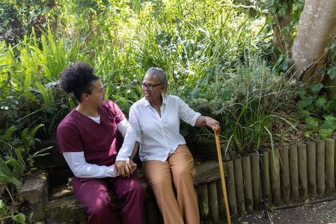Caregiver holding hands with elderly african american woman on garden bench, outdoor care