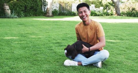 Smiling Asian Man Playing with Puppy on Sunny Green Lawn