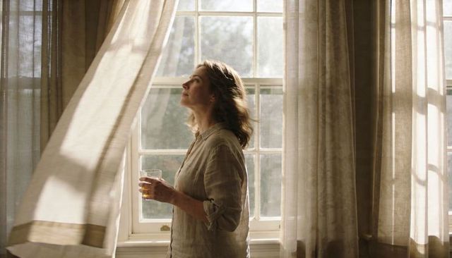 Contemplative woman holding amber glass by sunlit paned window with flowing curtains
