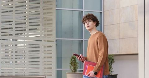 Young man holding smartphone and red folders near modern glass facade looking thoughtful