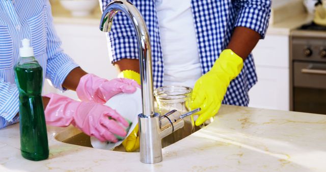 Two individuals are engaged in washing dishes at a kitchen sink, wearing protective gloves, with copy space. Their coordinated effort in maintaining cleanliness reflects a shared responsibility in household chores.