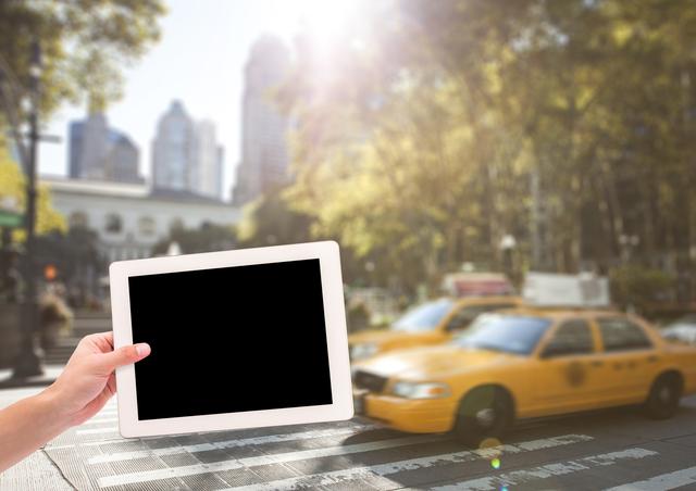 The image shows a hand holding a tablet in the foreground, with a blurred urban city background featuring a yellow taxi. This versatile visual could be used for promoting mobile apps, technology advancements, urban lifestyle, and transportation services. Its focus on the digital interface juxtaposed with daily city commute signifies modern connectivity.
