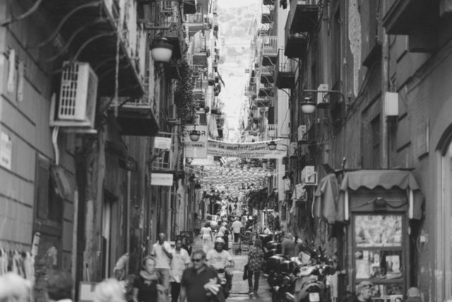 Image shows a narrow, busy alleyway in an old European city. Groups of people are walking, interacting, and going about their daily activities. Buildings on either side feature balconies, open windows, and multiple air conditioning units. Various signs and hanging lights add to the lively, historic atmosphere. This could be used for concepts related to European tourism, urban culture, historic towns, and lively cityscapes.