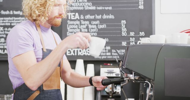 A male barista with curly hair preparing coffee in a modern cafe. He is using an espresso machine and holding a cup, focusing on his task. The cafe's menu board is visible in the background, suggesting a variety of offerings. This can be used for content related to coffee shops, barista training, customer service, or food industry settings.