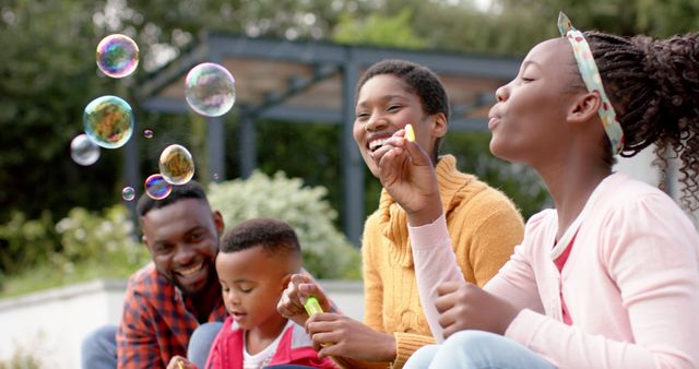 African American family of four enjoying quality time together in outdoor park. Parents and children blowing bubbles, smiling, and having fun. Ideal for concepts of family bonding, childhood enjoyment, parental love, leisure activities, and carefree moments.