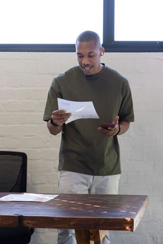 African american man standing at wooden desk reading documents while holding smartphone