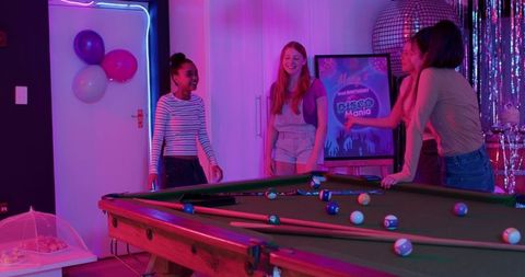 Young diverse women friends playing pool at neon disco birthday party with mirrorball and vibrant de