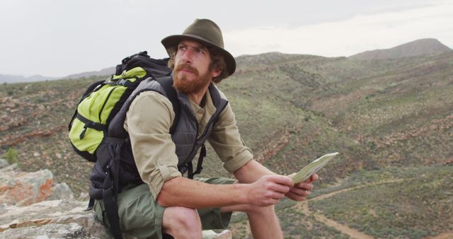 Bearded caucasian male survivalist sitting on mountain in wilderness, reading map and looking around. exploration, travel and adventure, survivalist in nature.