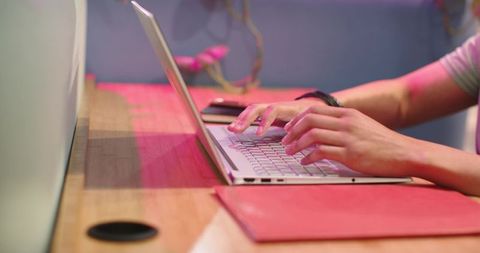 African man typing on silver laptop at wooden desk with smartphone, red folio, pink lighting