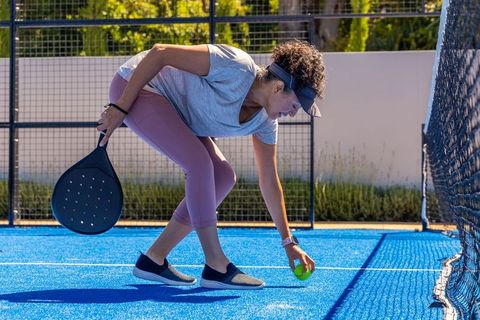African american woman picking paddle tennis ball on vibrant blue court under sunny sky