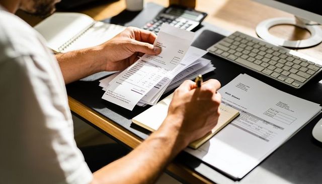 Man organizing receipts and writing notes at home desk with calculator for budgeting