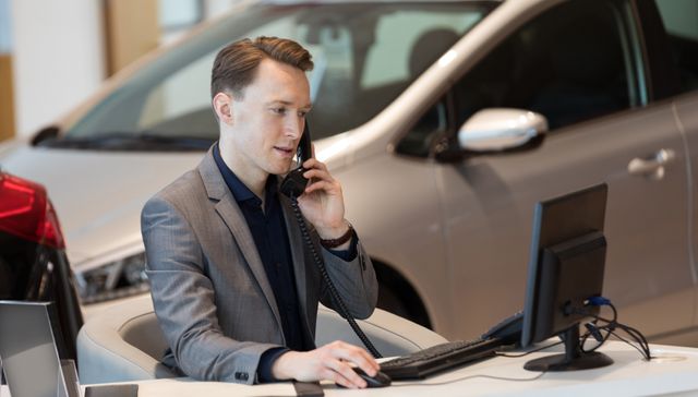 Salesperson talking on phone while using computer in car showroom. Ideal for illustrating business communication, customer service in automotive industry, and professional work environments. Useful for marketing materials, business presentations, and websites related to car dealerships and sales.