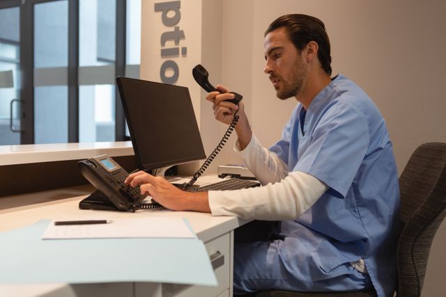 Male surgeon in blue scrubs dialing a landline telephone at a hospital reception desk. Ideal for use in healthcare, medical office, hospital administration, and communication themes.