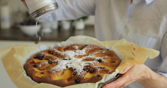 Person sprinkles powdered sugar over a freshly baked fruit tart in a home kitchen. Tart is topped with various fruits and appears freshly baked. Perfect for content relating to home baking, recipes, dessert preparation, or food blogs. Could be used in articles or features about homemade desserts, culinary skills, or baking techniques.