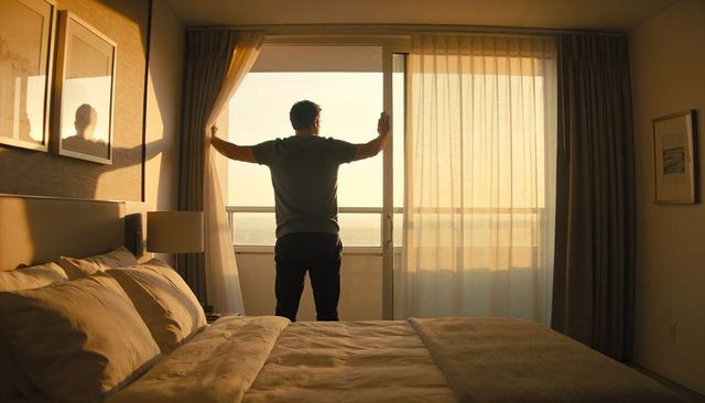 Man pulling aside sheer curtains at balcony door in sunlit hotel bedroom at sunrise