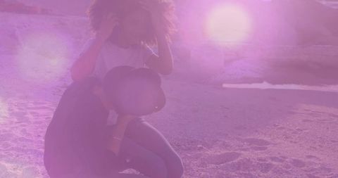Backlit couple crouching on sandy beach holding wide-brim hat with sun flare