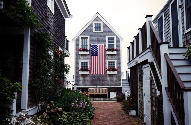 Traditional American house with gray siding features a large American flag and flower boxes on the upper windows. Brick walkway and wooden benches evoke a cozy and welcoming atmosphere, surrounded by other similar homes. Ideal for topics related to American patriotism, real estate, home decor, small-town living, and seasonal promotions.