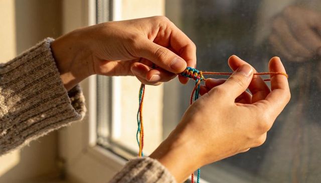 Close-up hands braiding colorful friendship bracelet with blue beads in sunlit window cozy