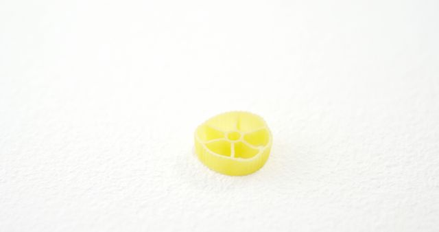 A single yellow pasta piece, resembling a small wheel or gear, is centered on a white textured background, with copy space. Its unique shape and bright color make it stand out, emphasizing the simplicity and beauty of everyday objects.