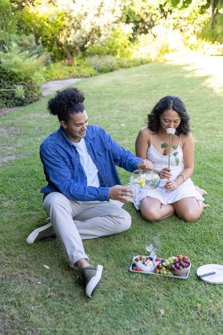 Couple enjoying picnic in garden, man pouring lemon water while woman smelling pink rose