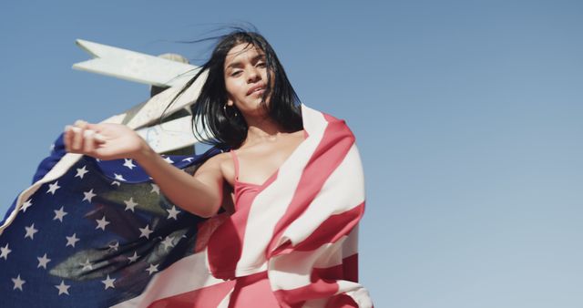 A young woman wearing a red dress is outdoors, celebrating with an American flag wrapped around her. She is looking at the camera with a relaxed nature and a clear blue sky in the background. This image can be used for patriotic events, summer holidays, advertisements for national pride, or social media posts promoting American independence and patriotism.