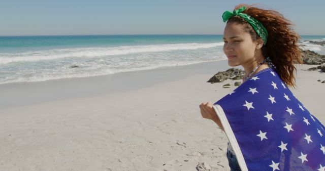 Young woman wrapped in American flag enjoying sunny day on sandy beach near ocean. Perfect for themes of patriotism, summer vacations, beach outings, national holidays like Independence Day, and freedom. Can be used in travel brochures, patriotic campaigns, lifestyle blogs, or advertisements emphasizing freedom and outdoor enjoyment.