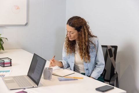 Mature woman working at minimalist conference table writing notes with laptop and coffee